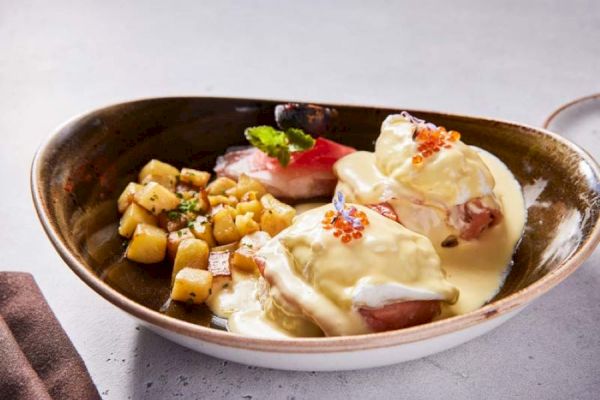 The image shows a plate with eggs Benedict topped with hollandaise sauce, garnished with herbs and served with diced potatoes.
