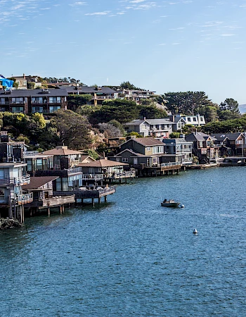 The image features waterfront houses situated along a hilly coastline, with a small boat in the water under a blue sky.