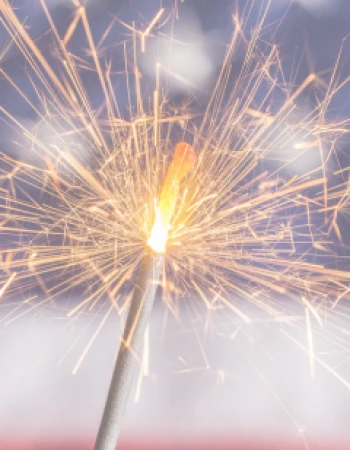A lit sparkler emits bright, colorful sparks in front of a blurred background resembling the American flag.